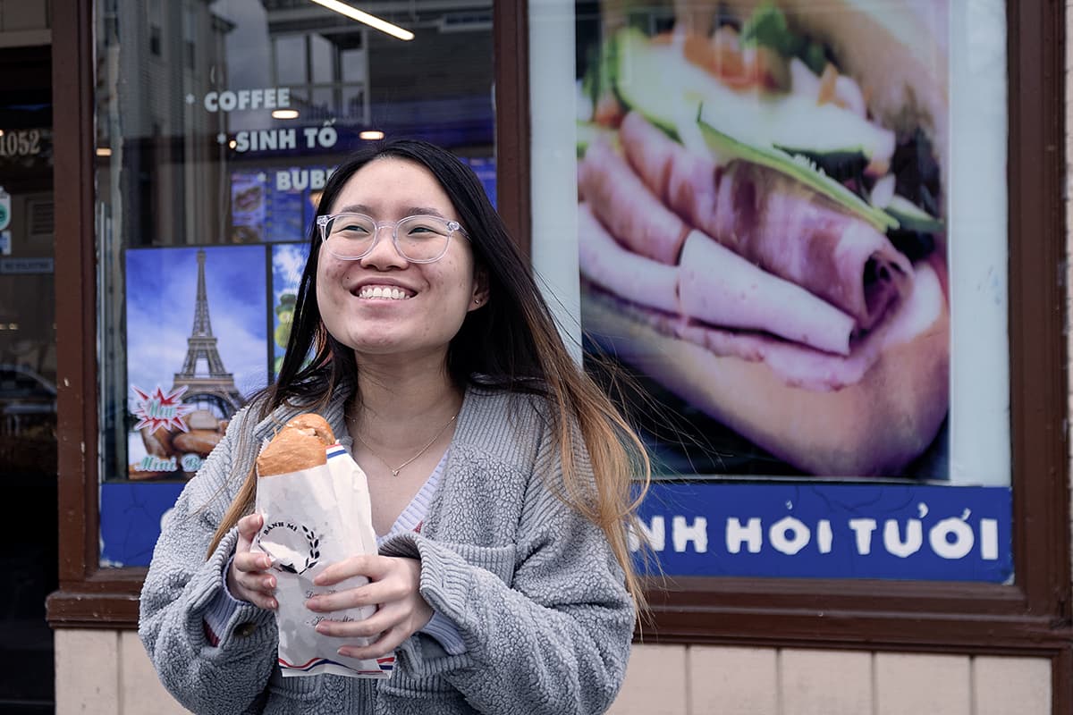 Student holds a banh mi sandwich in front of a shop in Dorchester.