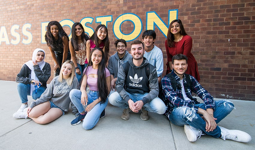 students seated on ground in a tight group in front of a UMass Boston sign on brick wall