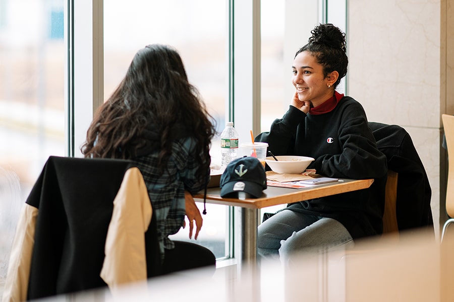 two female students talk casually at seated window in UMass Boston Campus Center Dining Hall