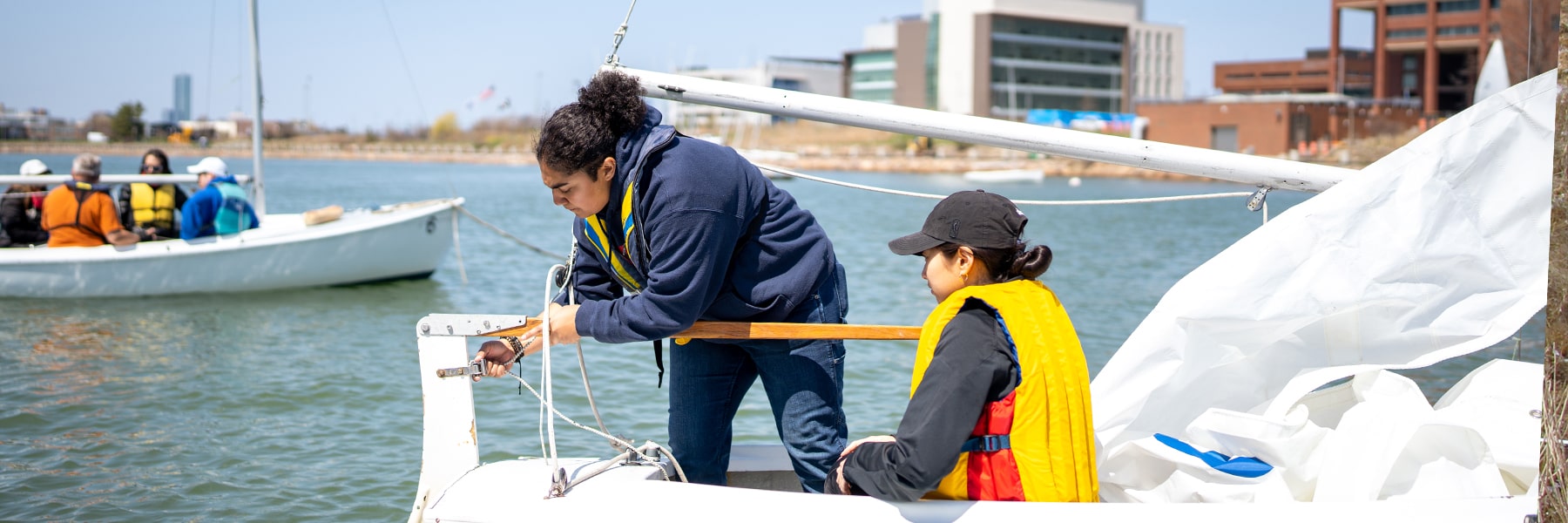 Two students work on sailboat in water near UMass Boston.