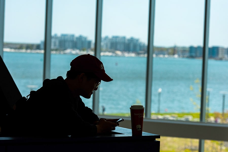Silhouette of student in baseball hat looks at cellphone next campus center window.
