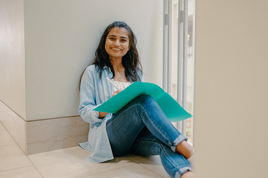 Nursing student sits on ground in campus center with laptop smiling.