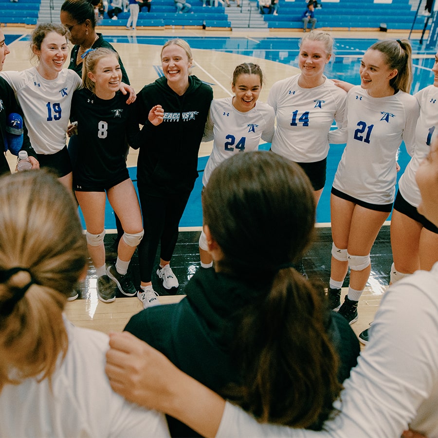 Girls volleyball players stand in circle on court smiling.