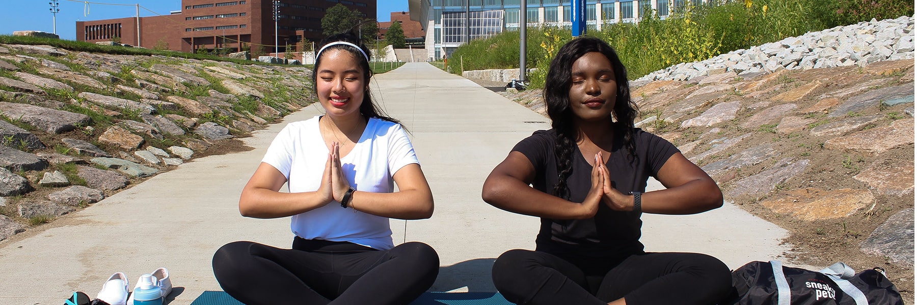 Two students sit crosslegged with hands together in a yoga pose in front of campus center.