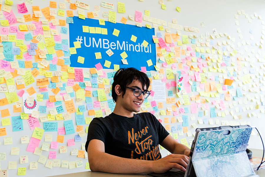 Student at campus center table with giant wall of stickies and prompt that states be mindful behind him.