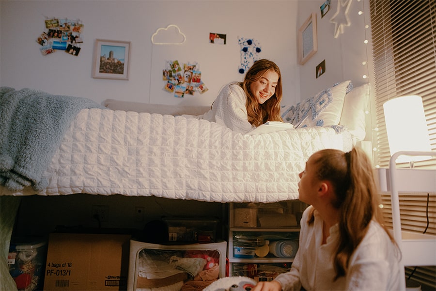 Two girls lounge on beds in dorm room.