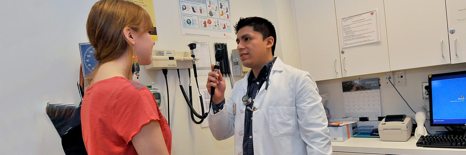 UMass Boston staff member in white coat with stethoscope examines student sitting on a medical table