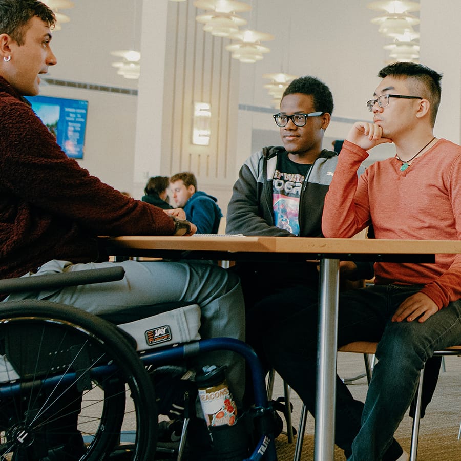 Stefan sits in wheelchair talks with two students in cafeteria.