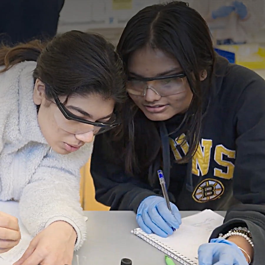 2 students in goggles looking closely together at research bench