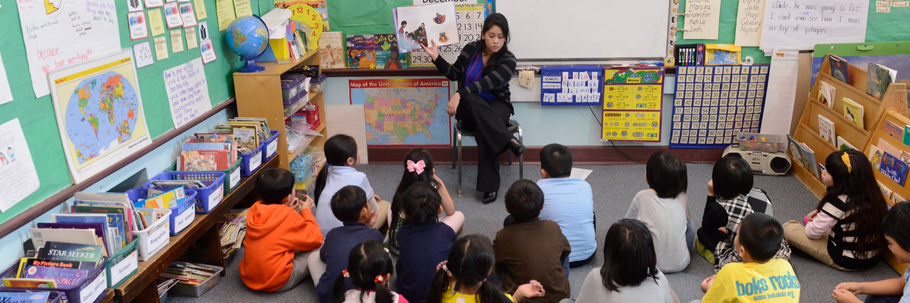 young teacher holding up a book and seated in front of large classroom of small children