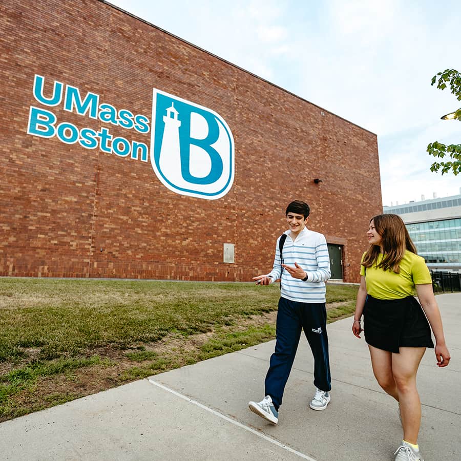 Students walking by brick wall with UMass Boston Logo