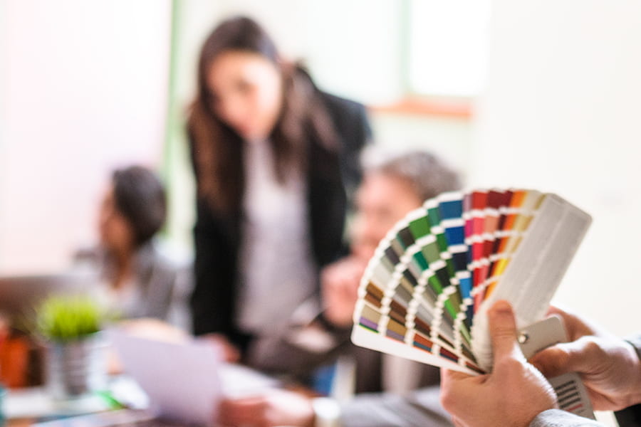 Designer holds pantone swatches while colleagues work in background.