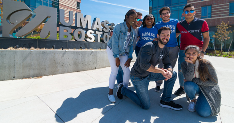 A diverse group of UMass Boston students in front of the sign outside the dorms 