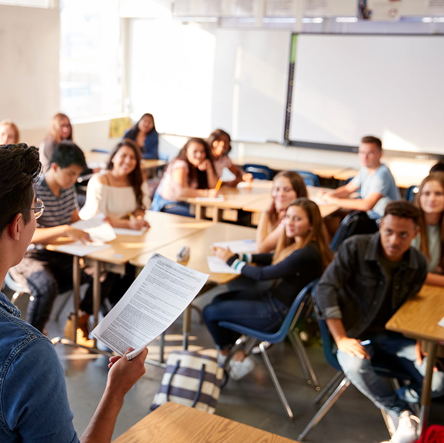 middle schoolers raising hands in class with back of teacher visible in front of classroom
