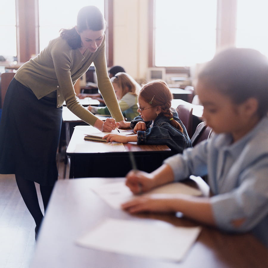 teacher bends over a child's school desk helping her out in the classroom