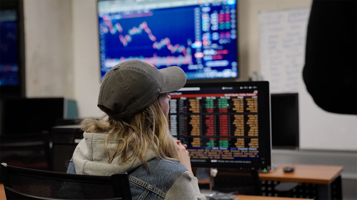 back view of student in the Bloomberg room in front of a large projected screen of candlestick charts on blue background and directly in front of student a computer screen of columns of data
