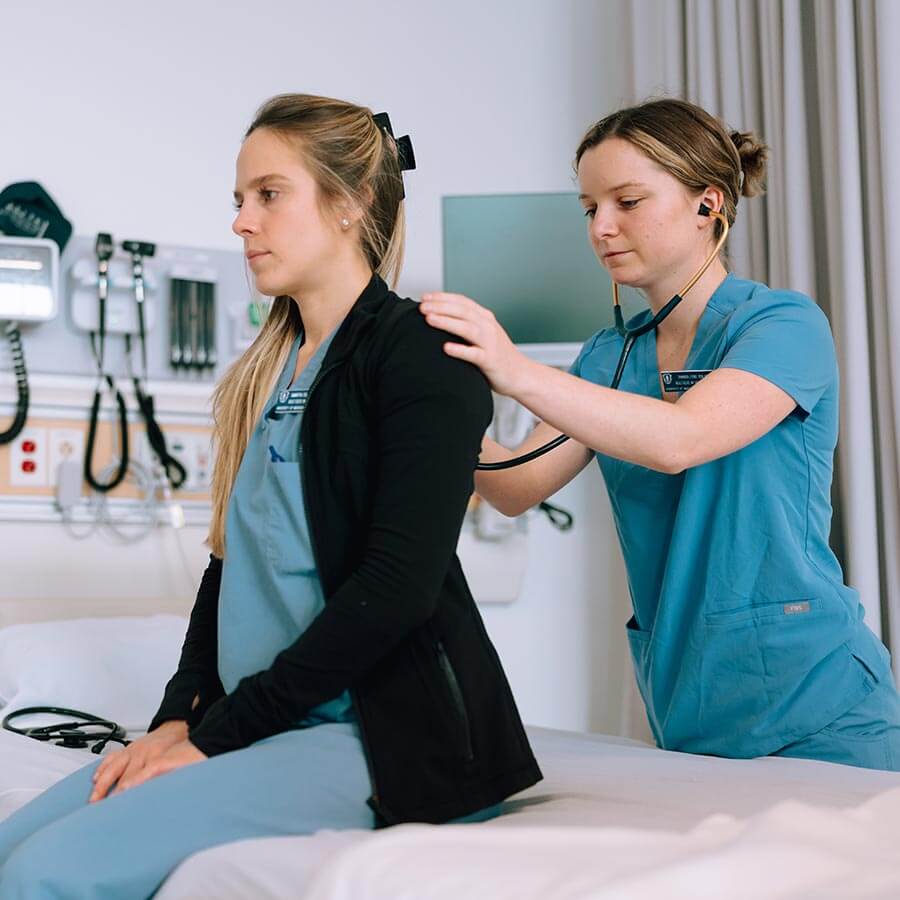 nursing student in light blue uniform holds stethescope to patient's back