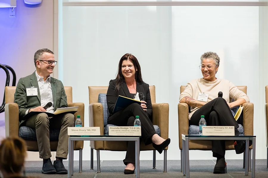 Three panelists seated onstage during a discussion event, smiling and holding microphones. Rhonda Hodge sits in the center between Steve Drury and Makeeba McCreary.