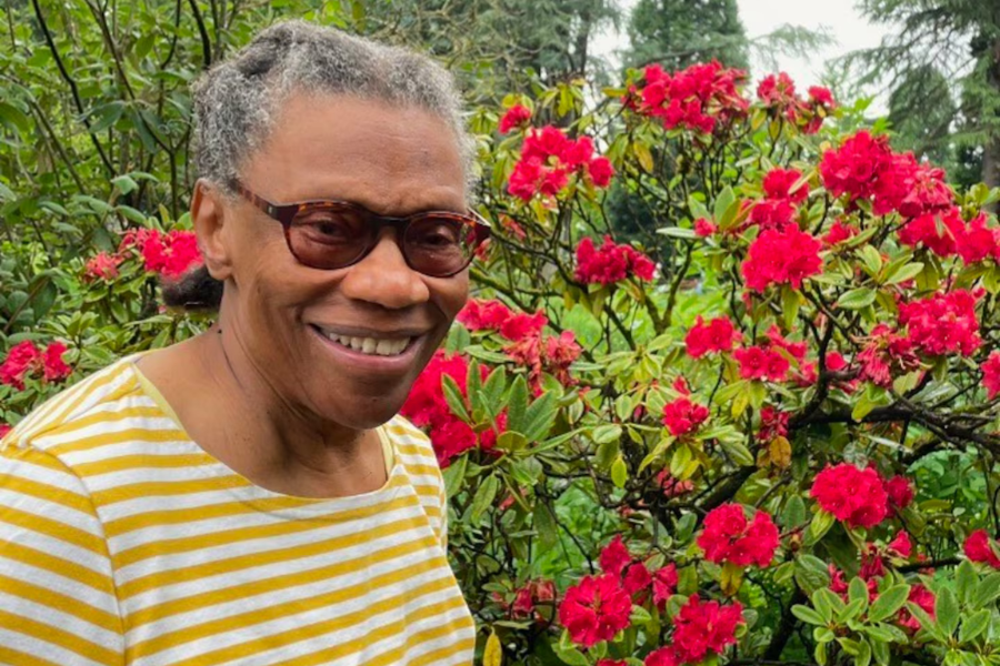 Photo of Boston State College alumna Joyce M. Grant smiling outdoors beside red flowering shrubs. She is wearing glasses and a yellow-and-white striped shirt, with lush greenery and blooming flowers in the background.