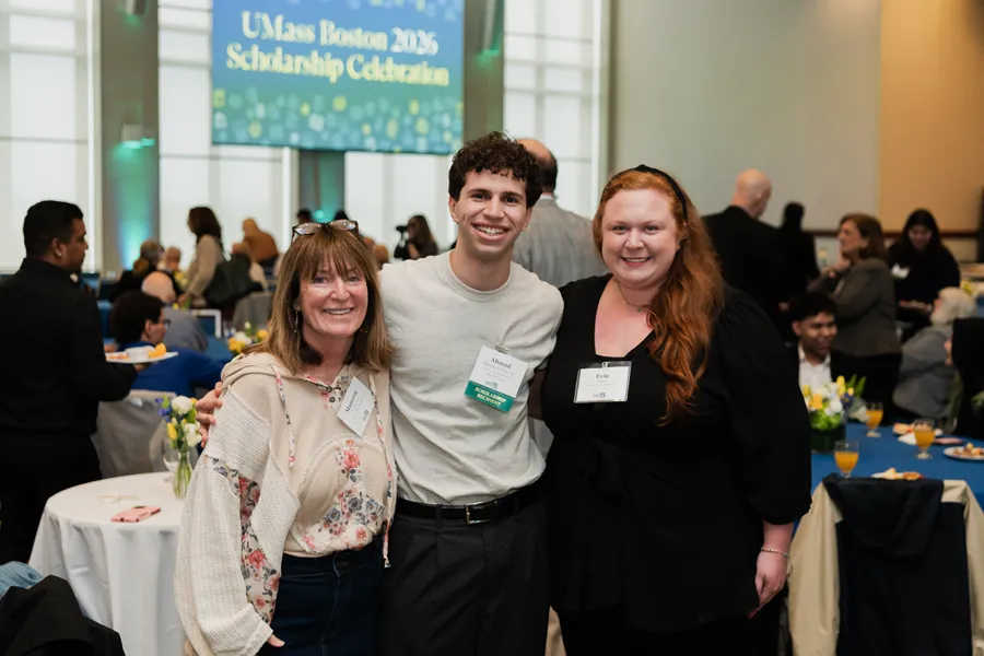 Three people smile together at a UMass Boston event with round tables and attendees in the background.