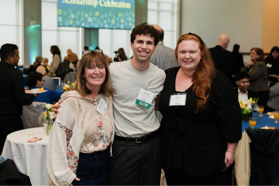 Three people smile together at a UMass Boston event with round tables and attendees in the background.