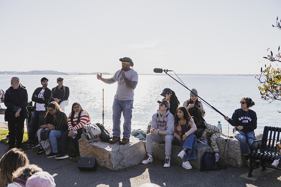 Faries Gray leading Indigenous Boston Harbor Tour, Indigenous man speaking to an audience on the shore of Deer Island, with the ocean in the background