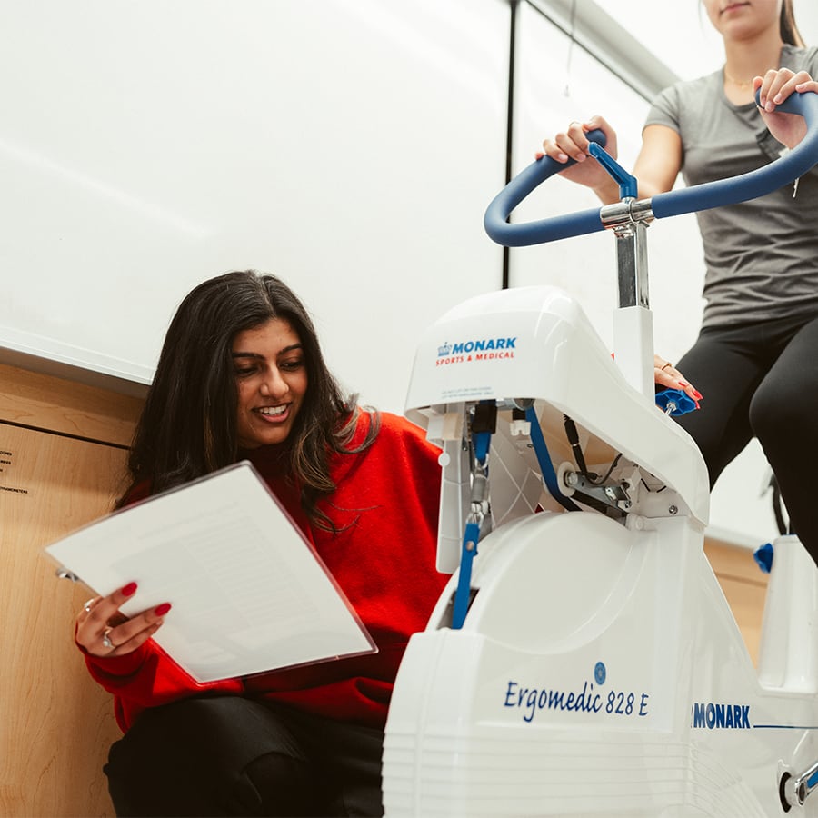 Student holding papers checks the wheel of exercise bike that has a rider.