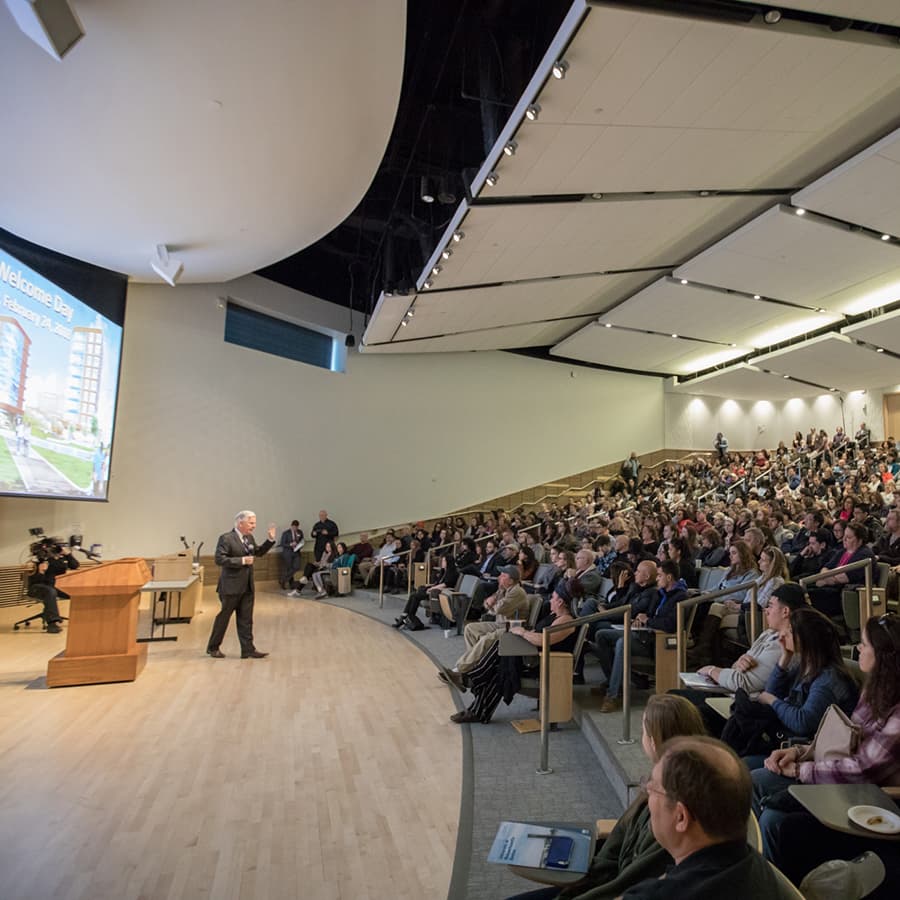 Presenter on stage in front of large screens speaking to audience in auditorium seating.