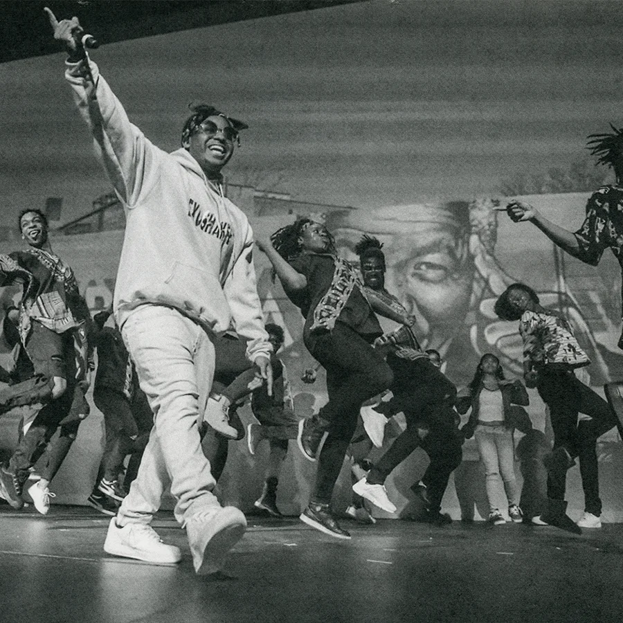A black and white photograph of a group of dancers on stage, smiling and with their hands in the air.
