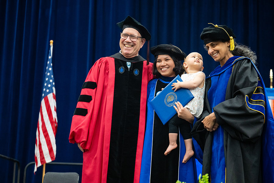 A doctoral degree recipient holds her baby, and her degree, onstage at the 2026 Doctoral Hooding ceremony