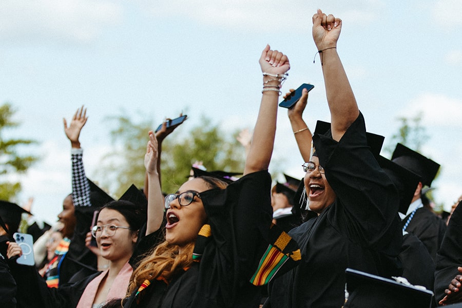 students in cap and gowns raise hands in celebration during commencement