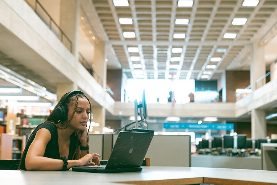 student wearing braids in library with headphones and laptop