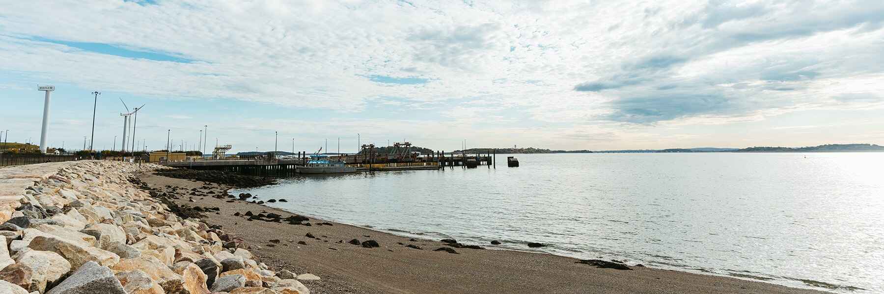 Rocks and sand along coastline with wind turbines, dock, and pier in the distance.