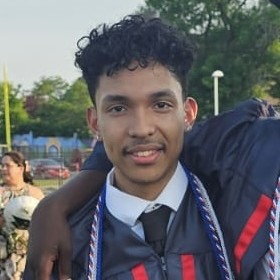 Young man with short, curly, dark hair looks into camera wearing graduation gown.